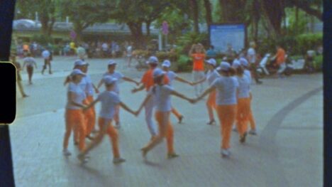 Still from a video work depicting a group of kids in uniform (composed of a white t-shirt and orange sweatpants) dancing in a circle on a public park.