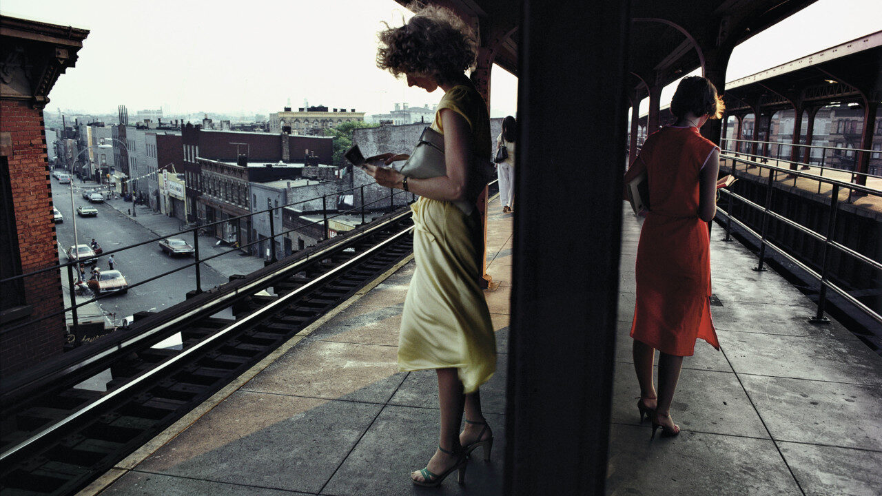 Photograph in colour of two women, wearing flowy dresses in contrasting yellow and red colours, standing in an overground subway platform in New York City. Their backs are turned to each other, and they are both reading a book.
