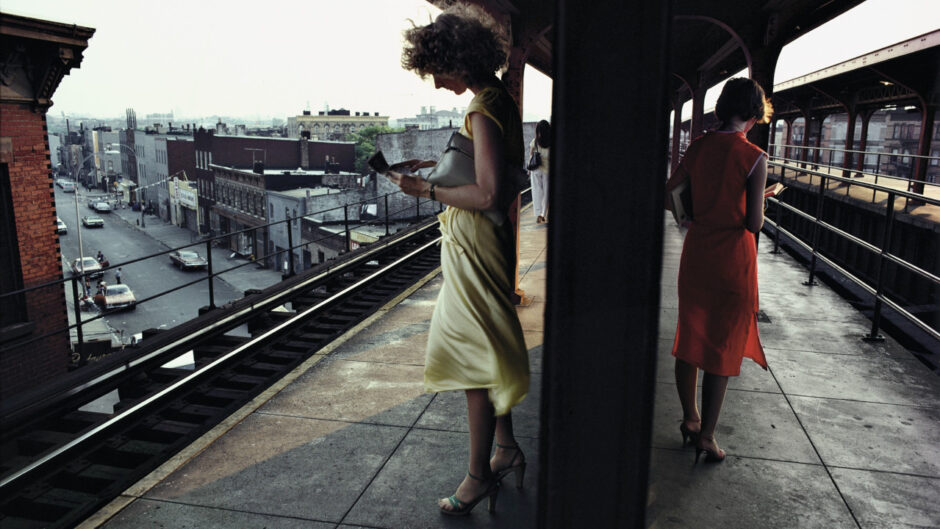 Photograph in colour of two women, wearing flowy dresses in contrasting yellow and red colours, standing in an overground subway platform in New York City. Their backs are turned to each other, and they are both reading a book.
