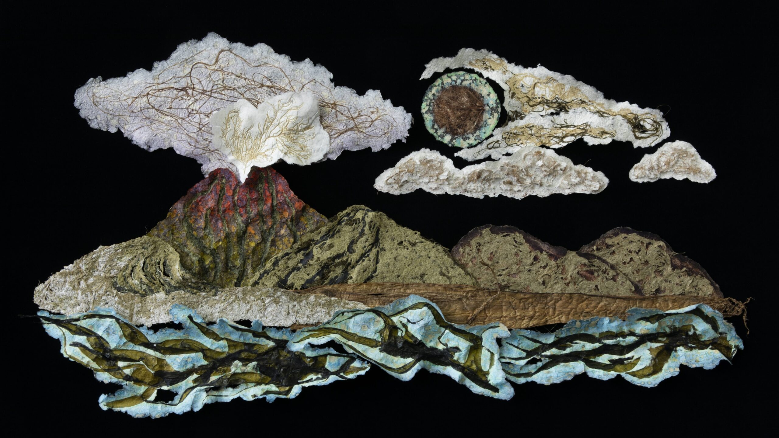 A photograph of a wall sculpture made of painted cardboard, depicting an island with an ignited volcano. The fumes from the volcano release into the sky, which is not rendered, acting as the negative space from the sculpture. There are cloud pieces floating above the large island piece, and a sun painted green.