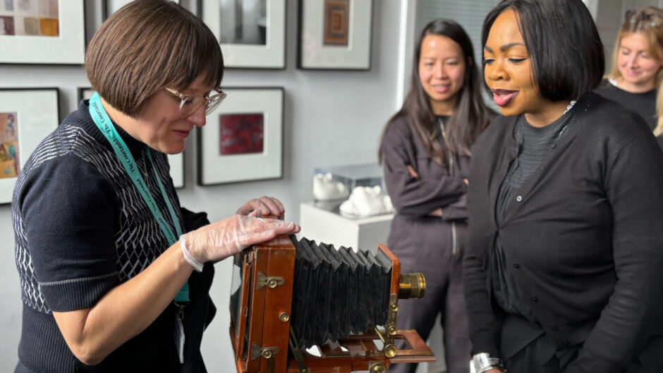 a group of women looking at an early photographic camera made of wood and metal.