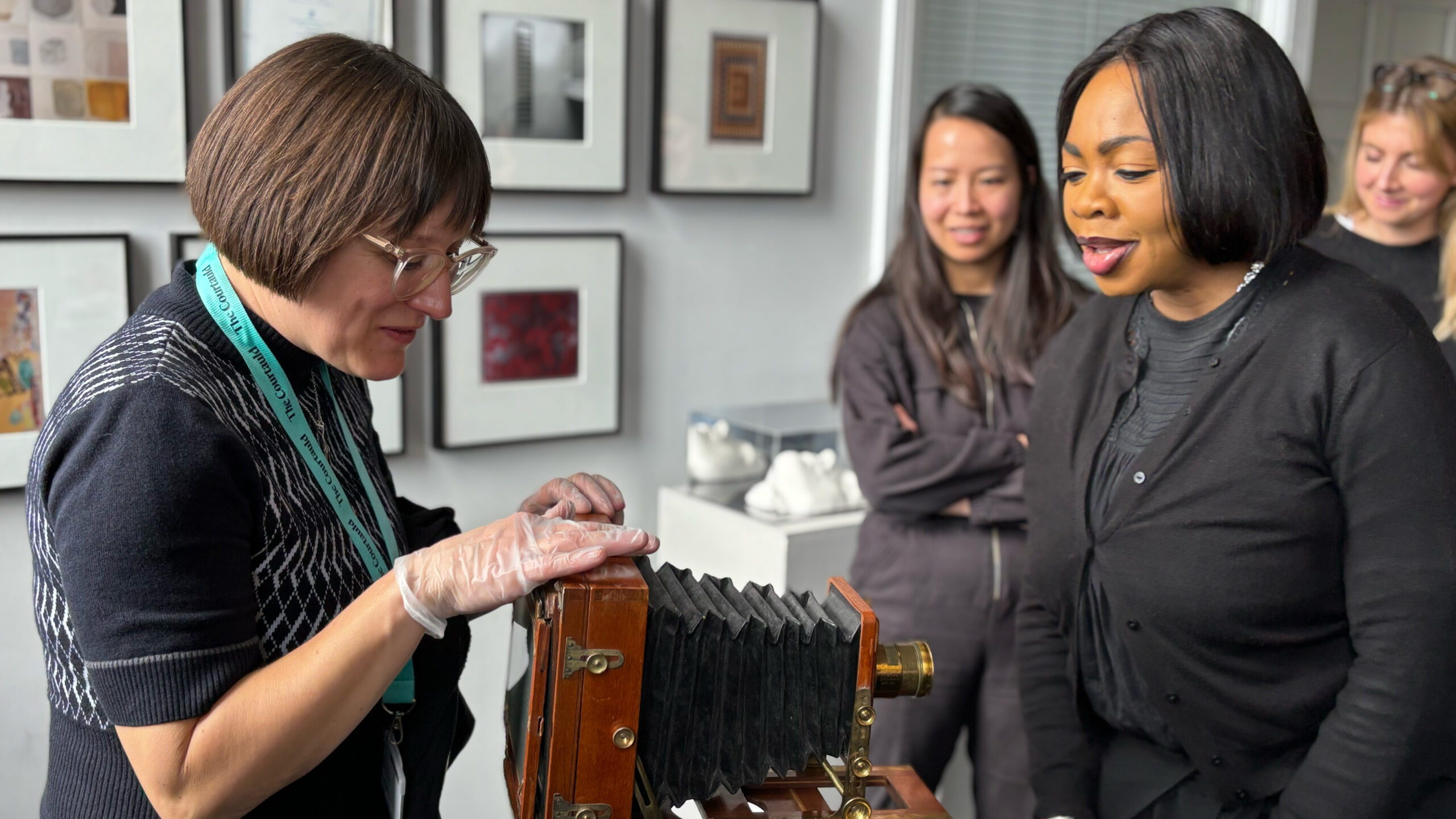 a group of women looking at an early photographic camera made of wood and metal.