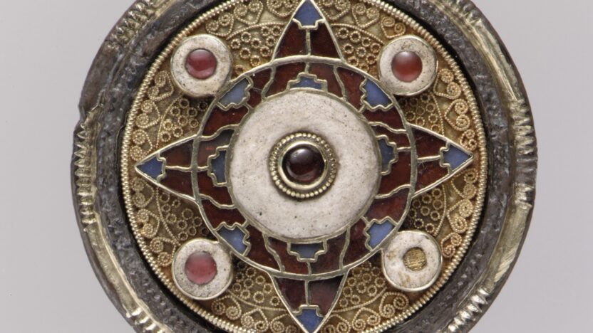 An early 600s disk-shaped brooch. Intricate traceries decorate the central part of the brooch, and four small jewels punctuate four corners of the disk. The shape of a cross is suggested in the centre of the brooch.