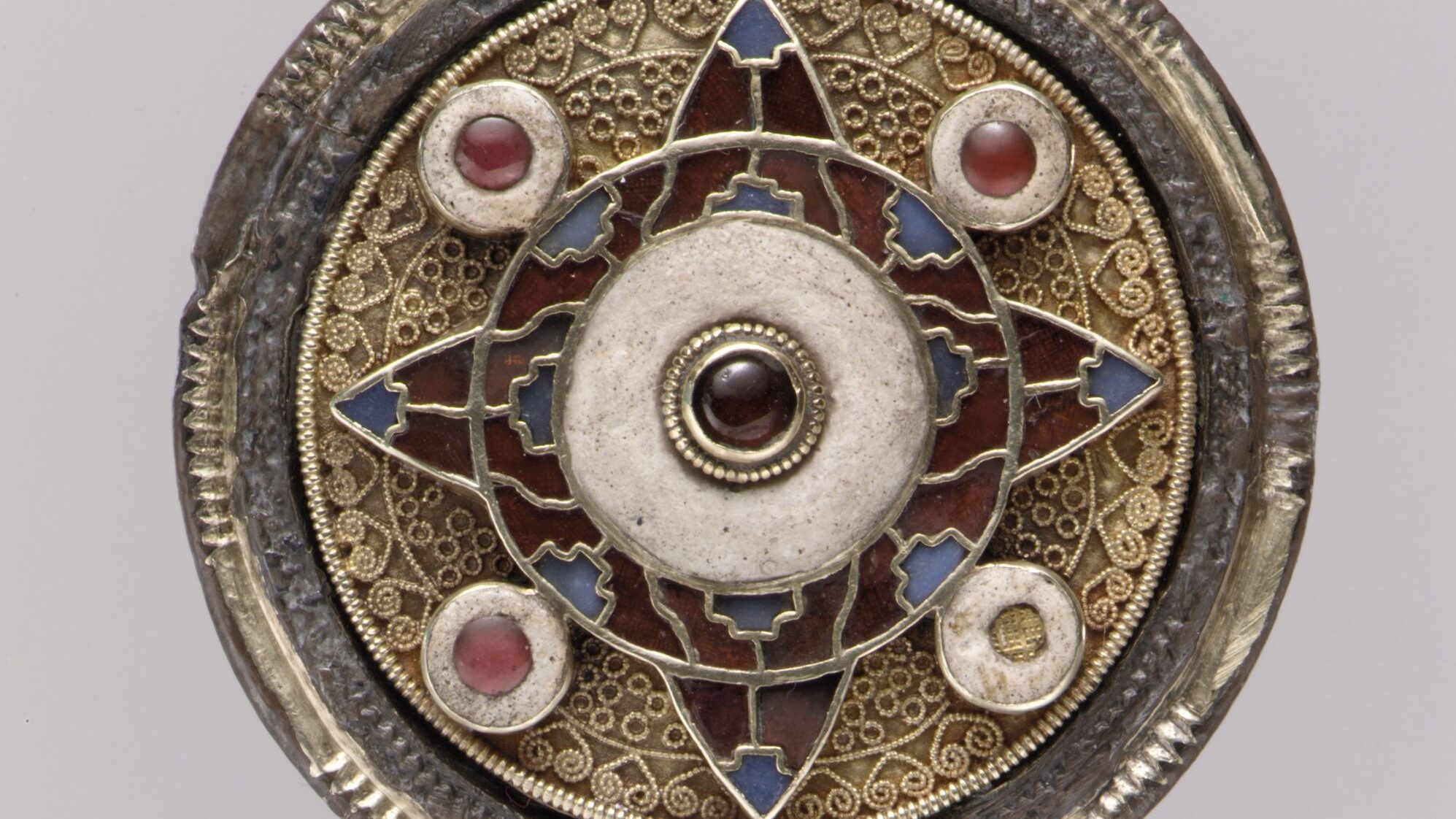 An early 600s disk-shaped brooch. Intricate traceries decorate the central part of the brooch, and four small jewels punctuate four corners of the disk. The shape of a cross is suggested in the centre of the brooch.