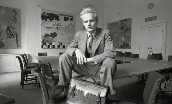 A blonde man sitting on the corner of a table looks defiantly at the camera. He is located in a university classroom decorated by large canvases with abstract art.