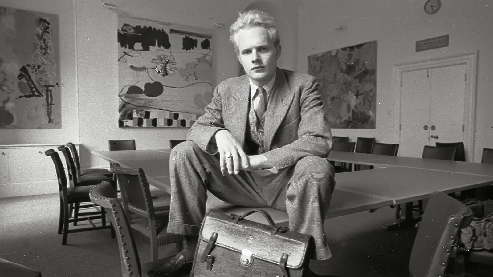 A blonde man sitting on the corner of a table looks defiantly at the camera. He is located in a university classroom decorated by large canvases with abstract art.