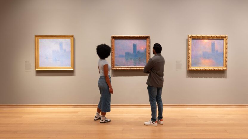 a man in a grey jacket and blue jeans, and a woman in a blue top and long denim skirt, both with dark hair, look at three of Monet's views of the Houses of Parliament on display at The Courtauld Gallery.