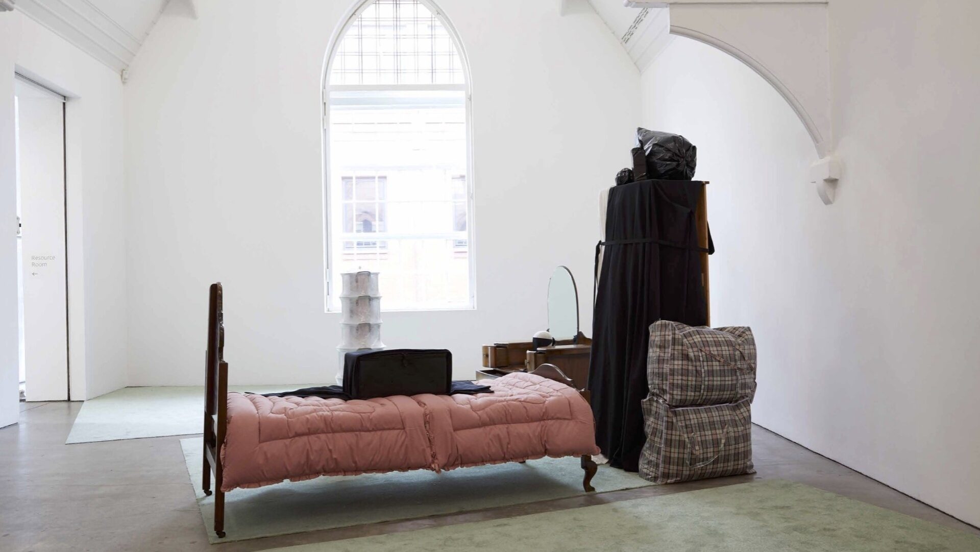 a white gallery space with a single dark wood bedframe and dressing table with several large bags filled with clothes and personal items
