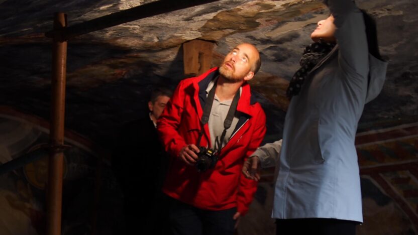 Professor Austin Nevin (a man in a red coat) and Professor Lori Wong (a woman in a blue coat) peer at a wall painting in a darkened room, illuminated by a flashlight.