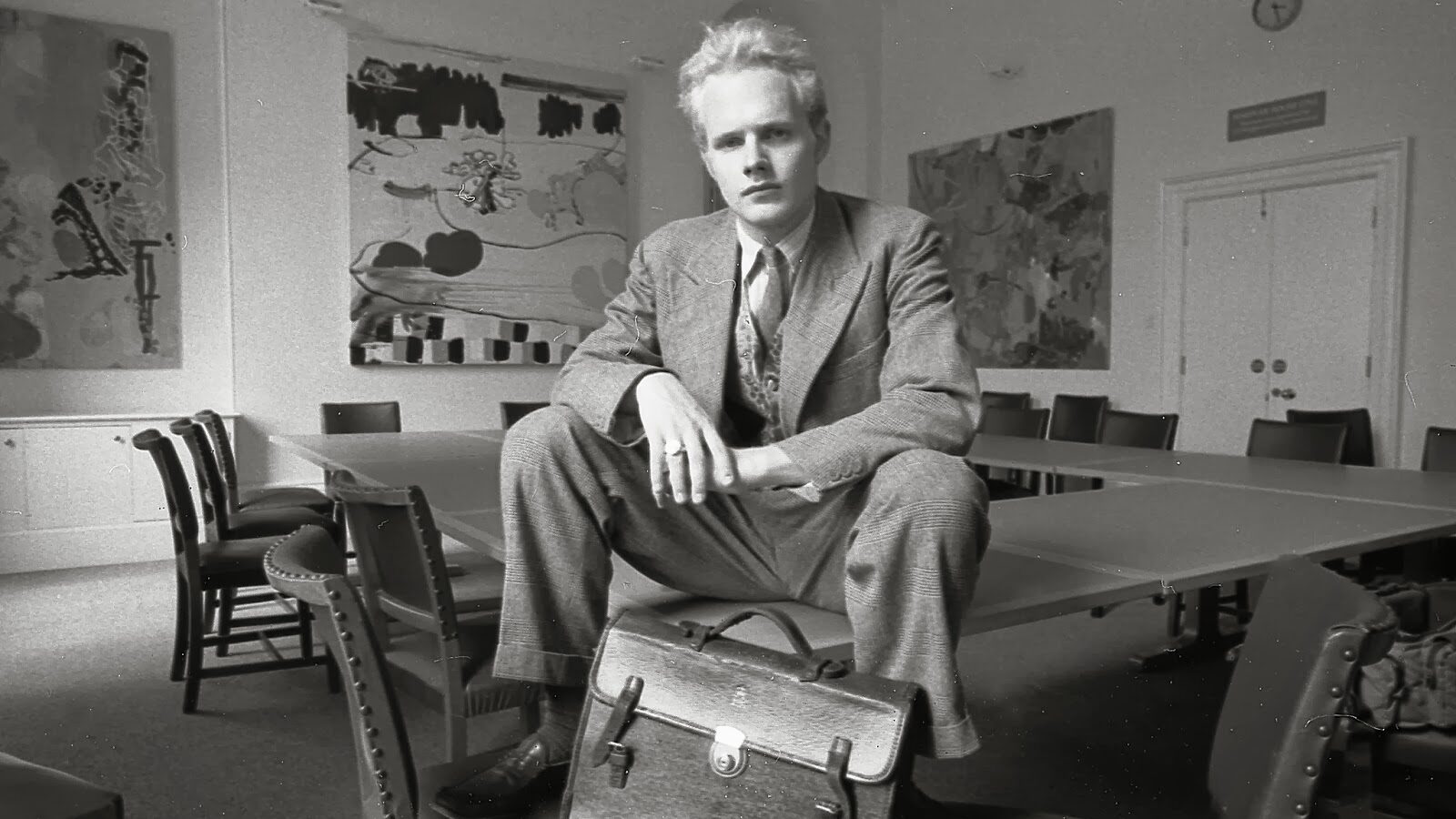A blonde man sitting on the corner of a table looks defiantly at the camera. He is located in a university classroom decorated by large canvases with abstract art.