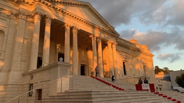 A large classical building with wide steps up to an imposing columned facade. The building, in Rome and in a light stone, is dappled in golden hour sunlight.