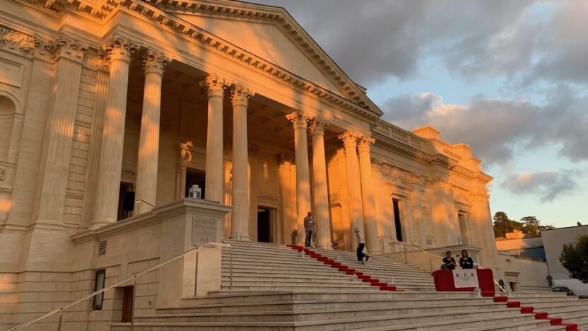 A large classical building with wide steps up to an imposing columned facade. The building, in Rome and in a light stone, is dappled in golden hour sunlight.