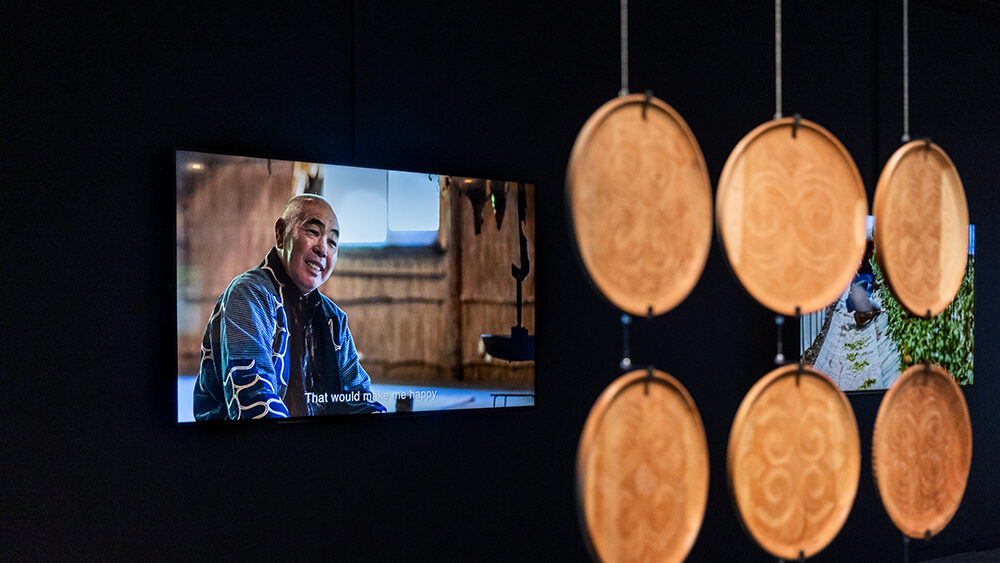 two screens displaying an interview with a member of the Biratori community play in the background of an art exhibition. Front right there is a hanging sculpture made of six wooden suspended discs in a. rectangular formation.