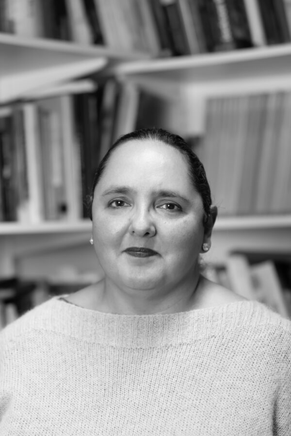 Black and white photograph of Prof Dorothy Price wearing a white jumper, smiling in an office with full bookshelves behind.