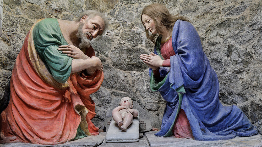 plaster sculptures of three biblical figures. Joseph in a orange and green robe far left, baby Jesus in a crib in the center an Mary in blue on the right.
