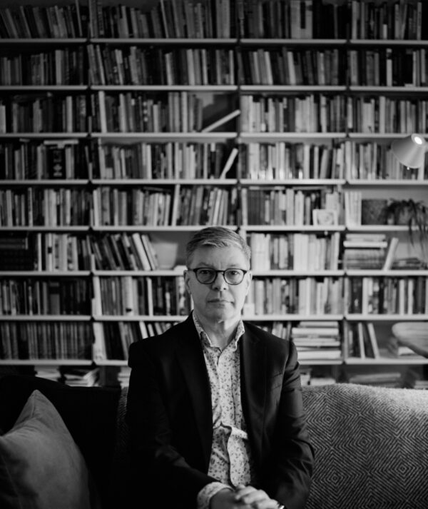 Black and white photograph of Steve Edwards wearing a patterned shirt and black blazer, with black glasses. He sits on a sofa in front of a floor-to-ceiling bookshelf full of books.
