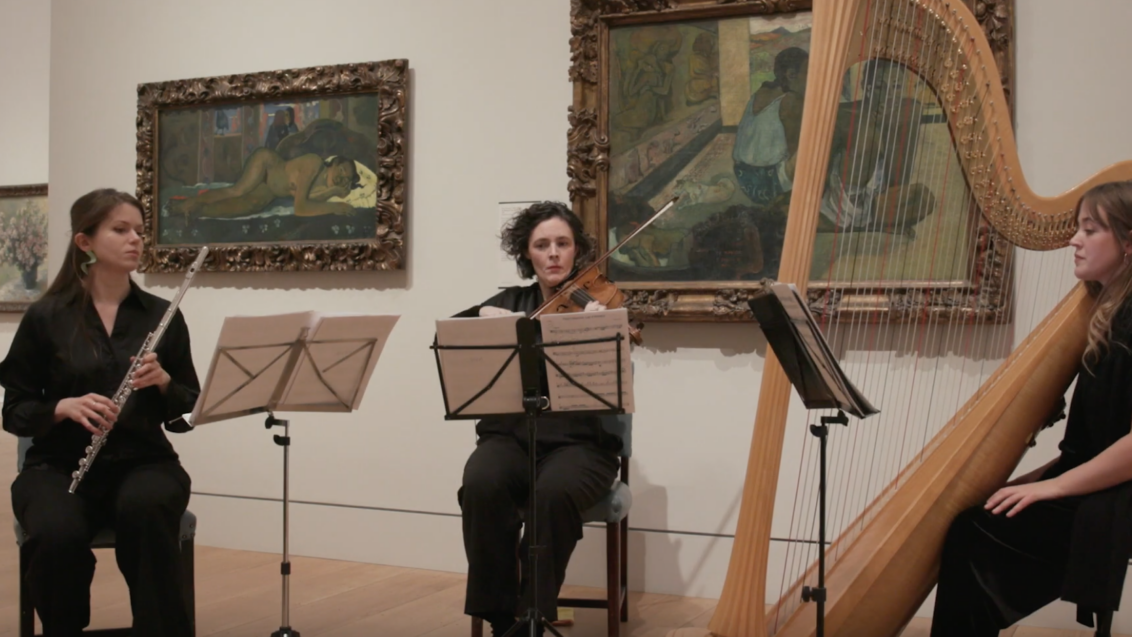 Three female students from the Guildhall School perform at the Courtauld Gallery in front of two Paul Gauguin paintings from his Polynesia series. On the left, a flute player is seated; in the center, a viola player is seated; and on the right, a harp player is seated.