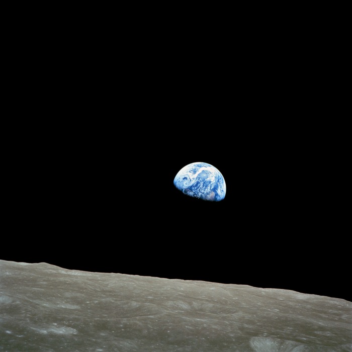 A photograph of Earth from lunar orbit, showing the small blue planet suspended in the vast darkness of space. The foreground features the Moon’s barren, beige surface, contrasting with the deep black sky and blue Earth.