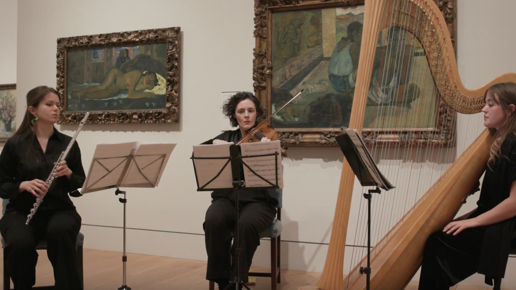 Three female students from the Guildhall School perform at the Courtauld Gallery in front of two Paul Gauguin paintings from his Polynesia series. On the left, a flute player is seated; in the center, a viola player is seated; and on the right, a harp player is seated.