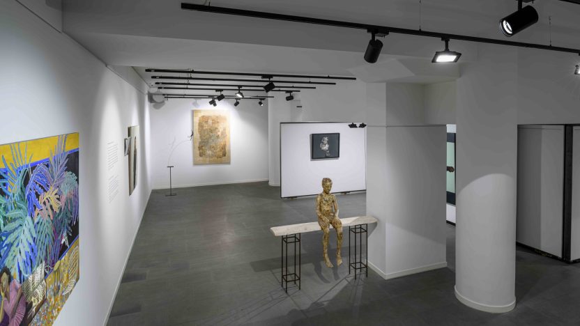 Panoramic shot of an exhibition installation, with prints and textiles visible on the wall and a sculpture of a human figure sat on a table most obviously in the centre.