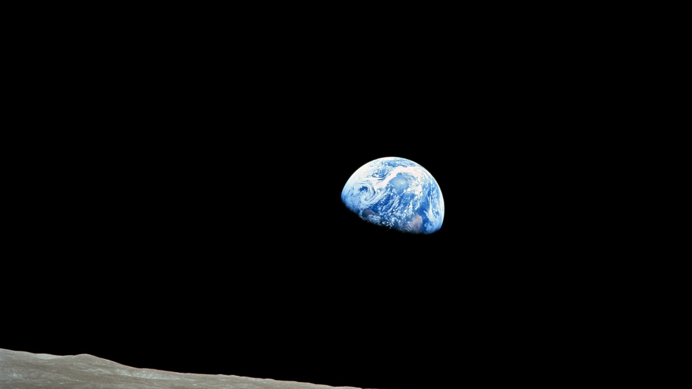 A photograph of Earth from lunar orbit, showing the small blue planet suspended in the vast darkness of space. The foreground features the Moon’s barren, beige surface, contrasting with the deep black sky and blue Earth.
