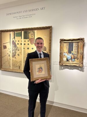 Konstantin Kandal standing in a Christie's auction house, smiling, and holding a small artwork framed in gold. He wears a dark suit and has a shaved head.