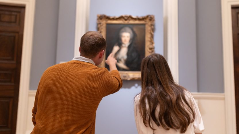 Two students sat on a bench, pictured from behind, pointing and looking at a painting by Thomas Gainsborough in the Blavatnik Fine Room.