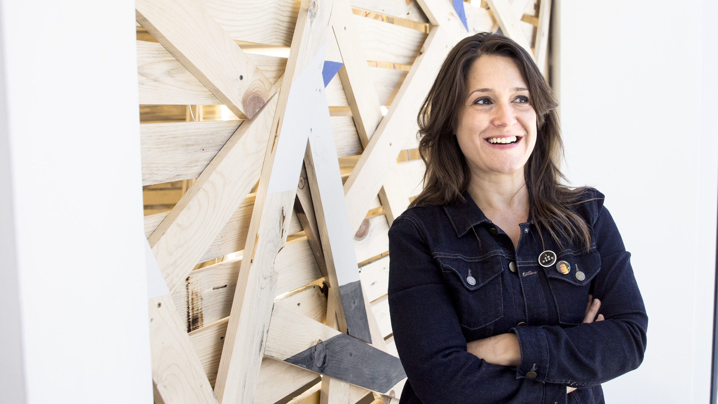 a woman stands smiling, dark shoulder length hair, pale skin, dark shirt.