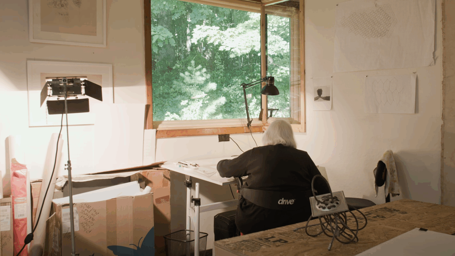 Alice Adams in her home studio, drawing at a large desk. She is surrounded by industrial-looking sculptures and is sat in a wheelchair.