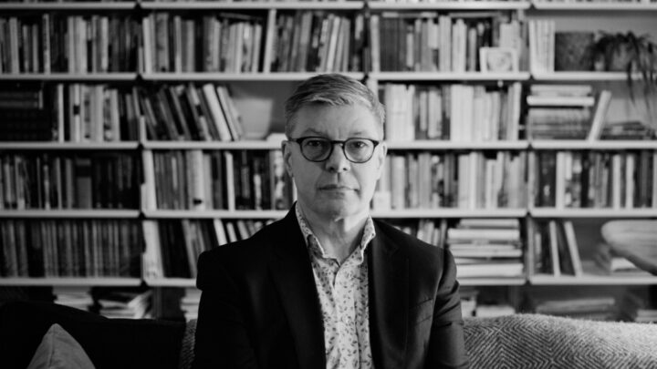 Black and white photograph of Steve Edwards wearing a patterned shirt and black blazer, with black glasses. He sits on a sofa in front of a floor-to-ceiling bookshelf full of books.