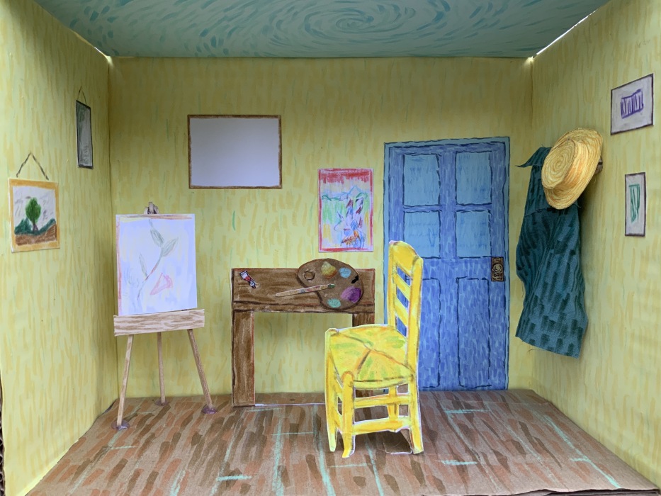 A handmade diorama of an artist’s room, inspired by Van Gogh’s style, features yellow walls, a blue door, and a wooden floor. It includes a yellow chair, easel, desk with art supplies, and framed artwork on the walls.