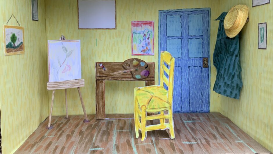 A handmade diorama of an artist’s room, inspired by Van Gogh’s style, features yellow walls, a blue door, and a wooden floor. It includes a yellow chair, easel, desk with art supplies, and framed artwork on the walls.