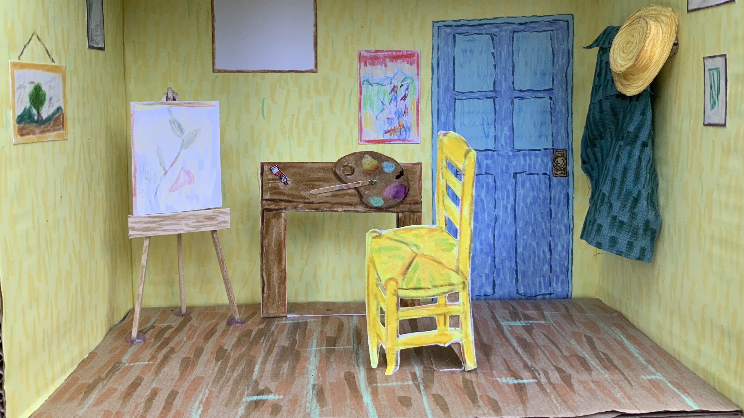 A handmade diorama of an artist’s room, inspired by Van Gogh’s style, features yellow walls, a blue door, and a wooden floor. It includes a yellow chair, easel, desk with art supplies, and framed artwork on the walls.