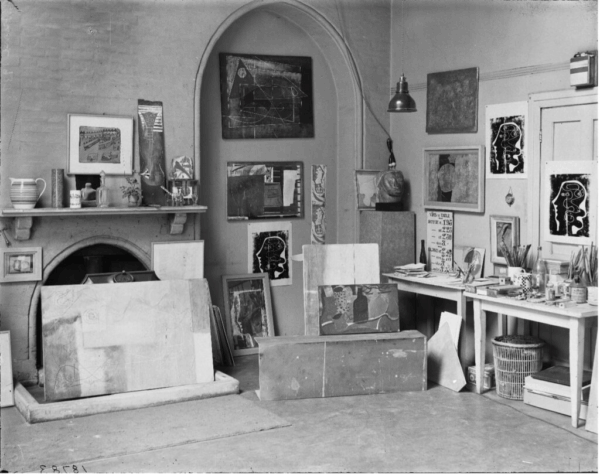 A photograph of an artist studio, with canvases and materials stacked against the walls and floor. Black and white photo.