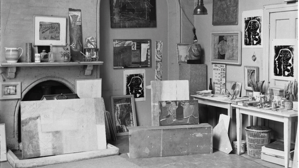 A photograph of an artist studio, with canvases and materials stacked against the walls and floor. Black and white photo.