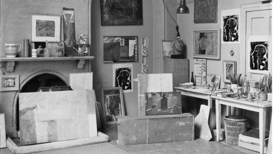 A photograph of an artist studio, with canvases and materials stacked against the walls and floor. Black and white photo.
