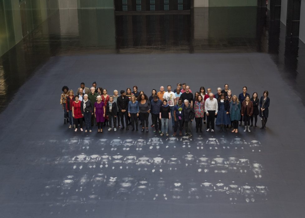 White paint body prints are spread across a grey floor in a large room, with a group of people standing behind them.