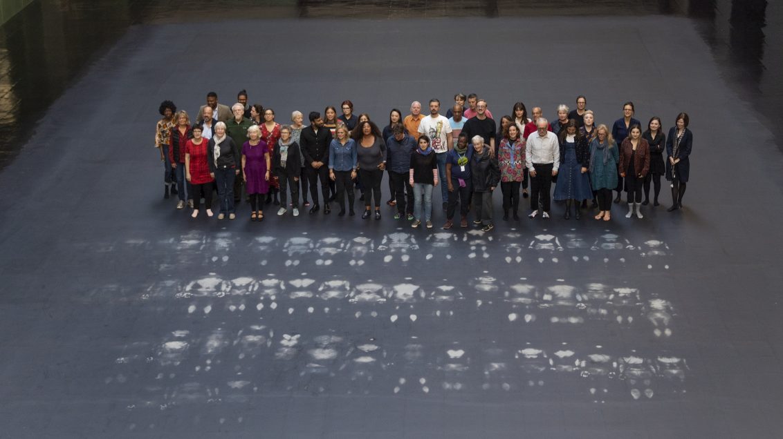 White paint body prints are spread across a grey floor in a large room, with a group of people standing behind them.