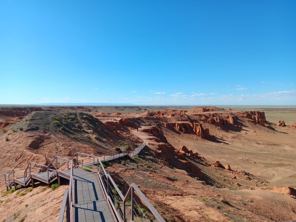 a decked walkway stretches out over a red desert like landscape with a bright blue sky.