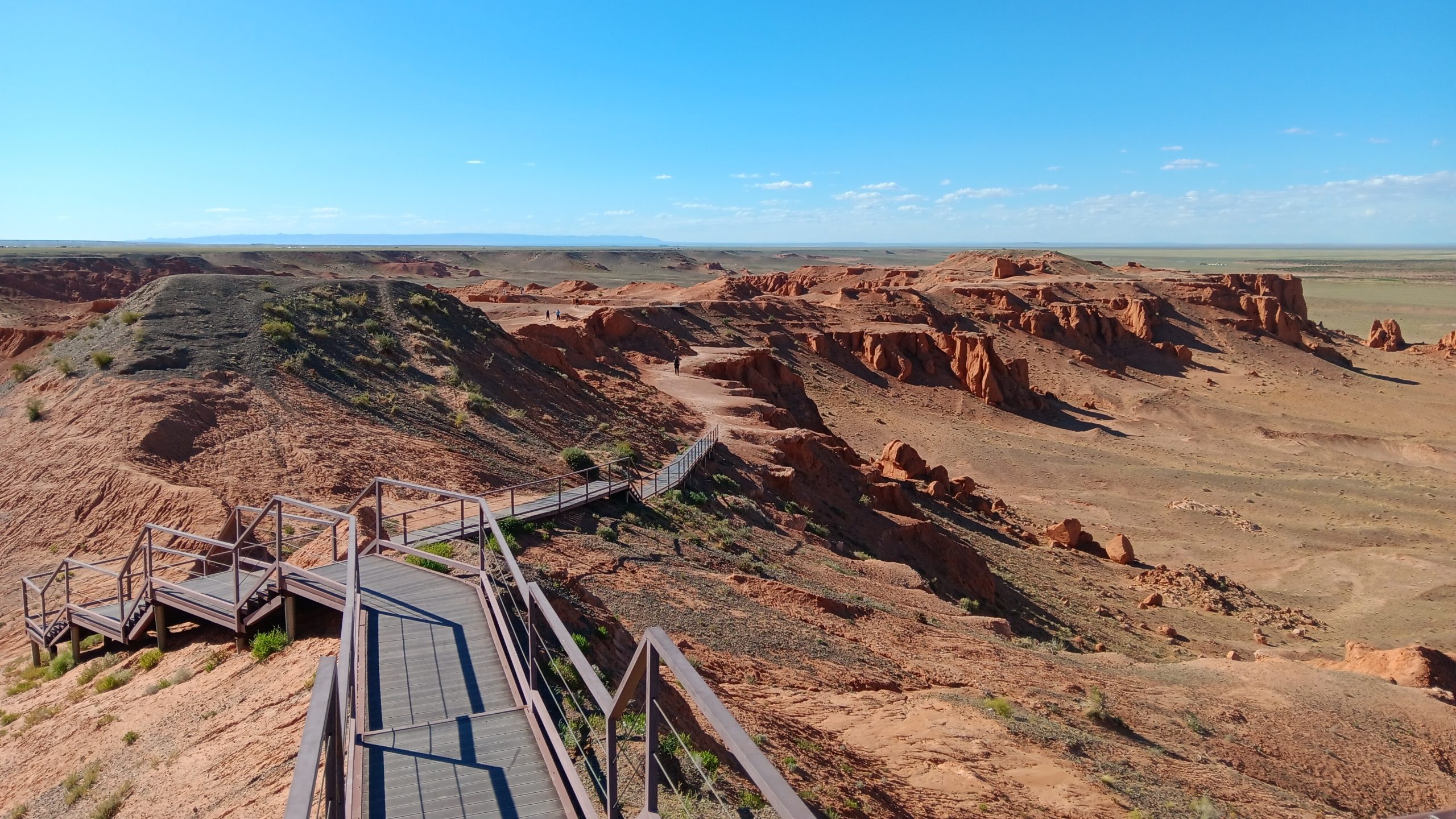 a decked walkway stretches out over a red desert like landscape with a bright blue sky.