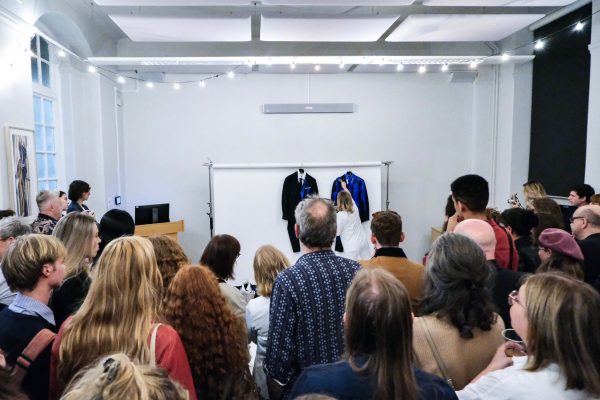 A group of people watching a performance with two suits hung against a white backdrop, in a classroom setting.
