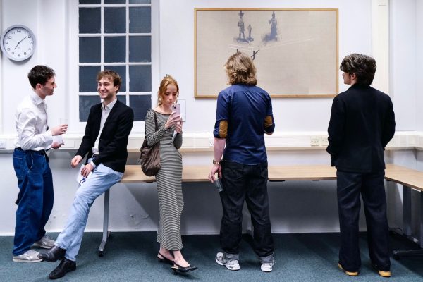 Four people sit/stand on tables in a classroom, chatting and looking at a large abstract artwork on the wall.