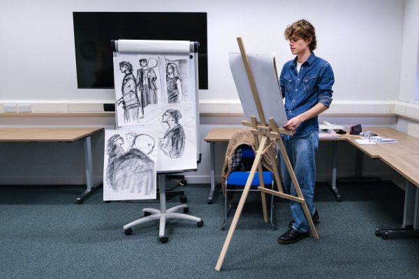 A young, male artist at an easel creates live sketches of the opening night. Two paper sketches are visible next to him, with drawings of people at the opening night.