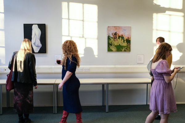 Three people talk and look at the artworks on the wall, as sunlight streams through a large window.