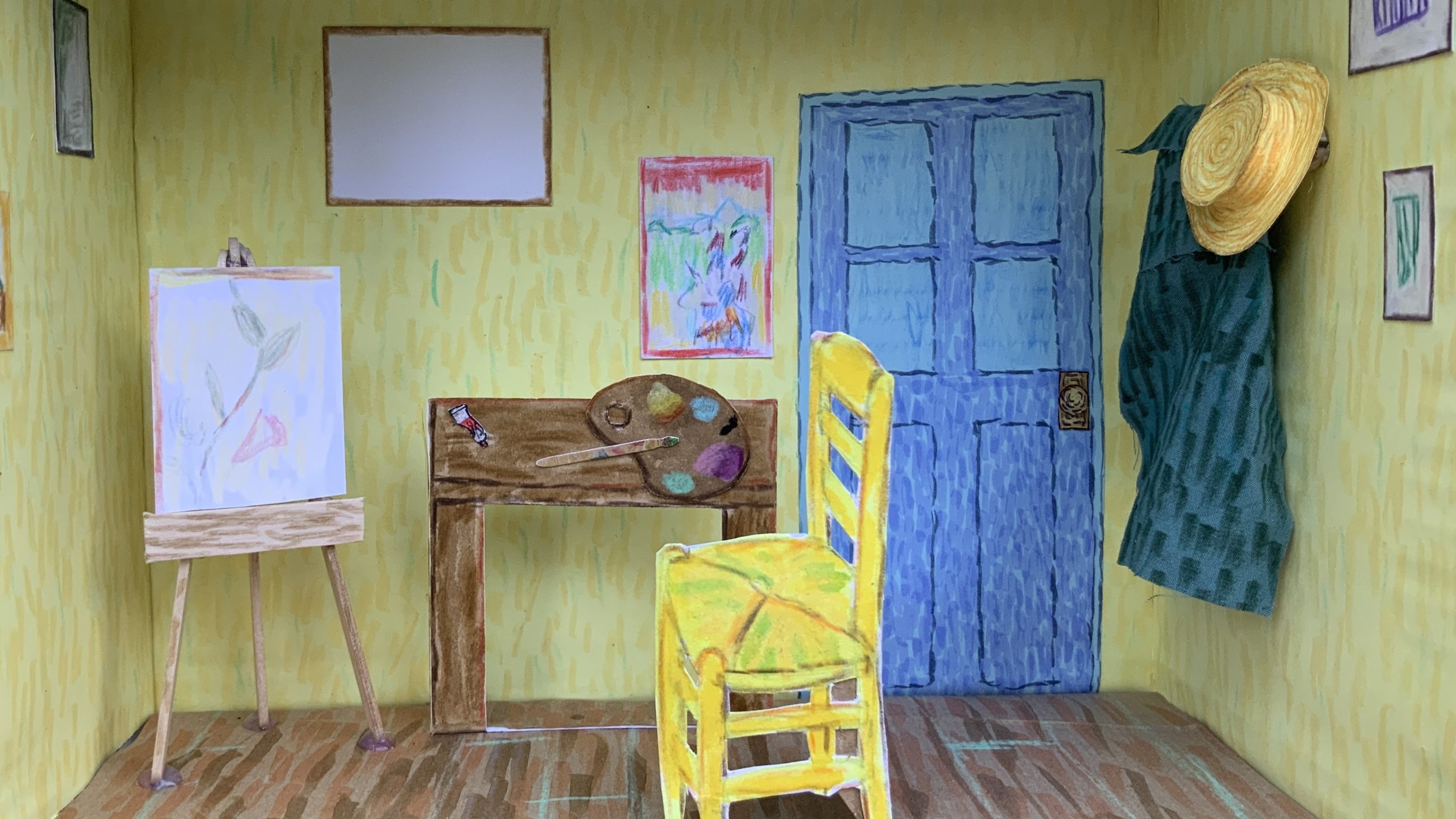 A handmade diorama of an artist’s room, inspired by Van Gogh’s style, features yellow walls, a blue door, and a wooden floor. It includes a yellow chair, easel, desk with art supplies, and framed artwork on the walls.