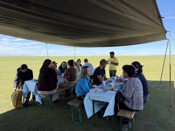 A large group sit outdoors across two tables under tents in the Mongolian landscape