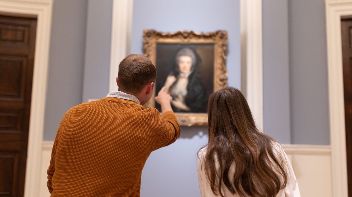 Two students sat on a bench, pictured from behind, pointing and looking at a painting by Thomas Gainsborough in the Blavatnik Fine Room.