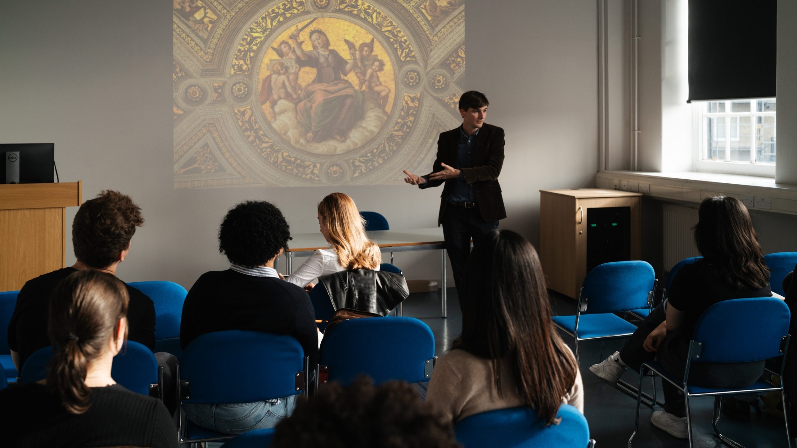 a lecturer in a seminar room pointing at an image projected on a wall, he's talking in front of a class of students that are looking towards him