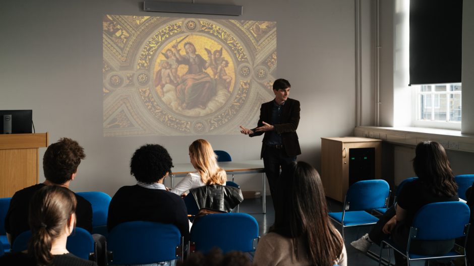a lecturer in a seminar room pointing at an image projected on a wall, he's talking in front of a class of students that are looking towards him