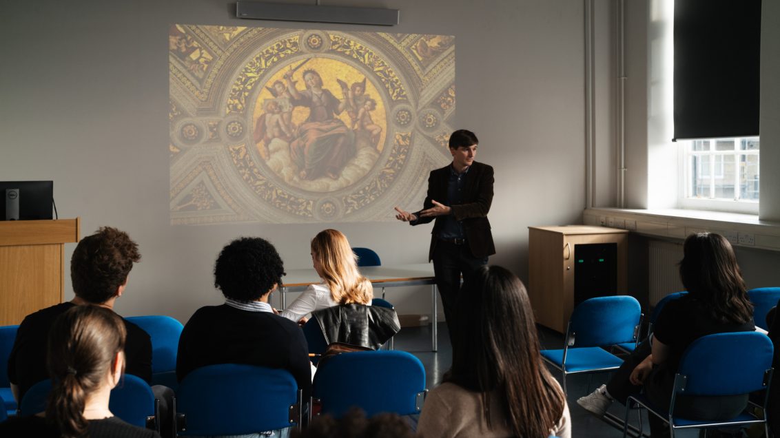a lecturer in a seminar room pointing at an image projected on a wall, he's talking in front of a class of students that are looking towards him